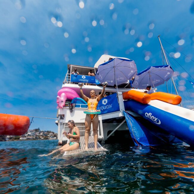 People enjoying a sunny day on a boat with a slide. One person stands at the end of the slide with arms raised, wearing a swim cap and goggles. Another person in a colorful swimsuit descends the slide. Inflatables and umbrellas are visible.