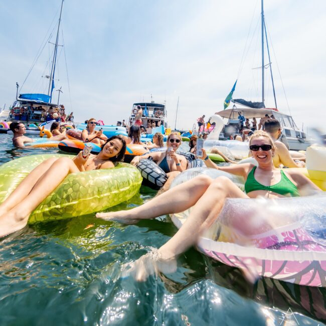 A group of people enjoying a sunny day on inflatable rafts in the water, surrounded by boats. They are smiling and relaxing. The background features several anchored yachts under a clear sky.