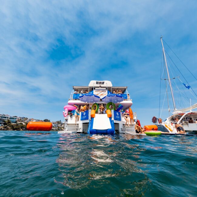A lively yacht party with people enjoying the sunny day. Inflatable toys, colorful objects, and swimmers surround the boat. Another yacht is nearby, and people are socializing in and out of the water under a blue sky.