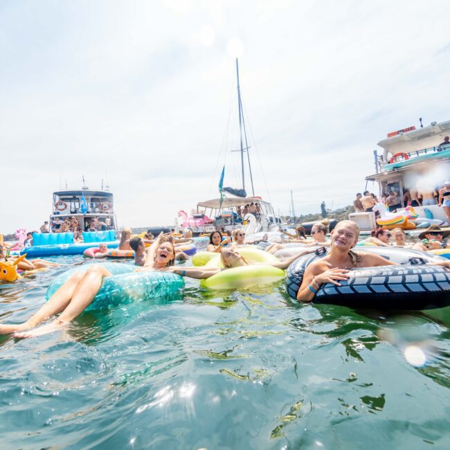 People enjoying a sunny day in the water surrounded by boats. Some are relaxing on colorful inflatables. The atmosphere is lively and festive, with boats in the background.