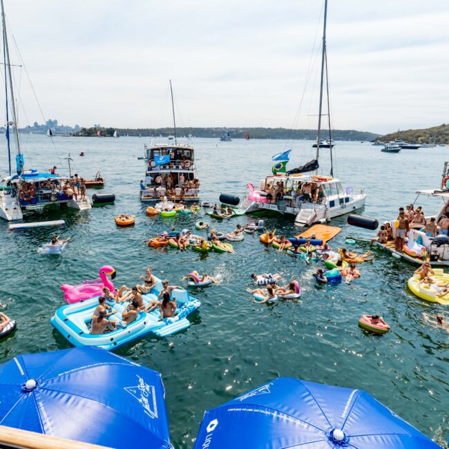 A festive gathering of people on a sunny day with colorful inflatables in the water between anchored boats. Participants are enjoying the water, with near and distant landscapes, and a bridge visible in the background.