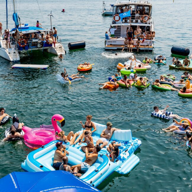 People relax on inflatable pool floats in a lake, surrounded by anchored boats. The scene is festive, with a variety of colorful floats including a pink flamingo and a blue unicorn. Sailboats and a distant skyline are visible in the background.