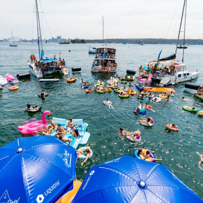 A lively scene with numerous people on colorful inflatables floating between several anchored boats in a bay. Two large blue umbrellas are in the foreground, and a city skyline can be seen in the distant background.