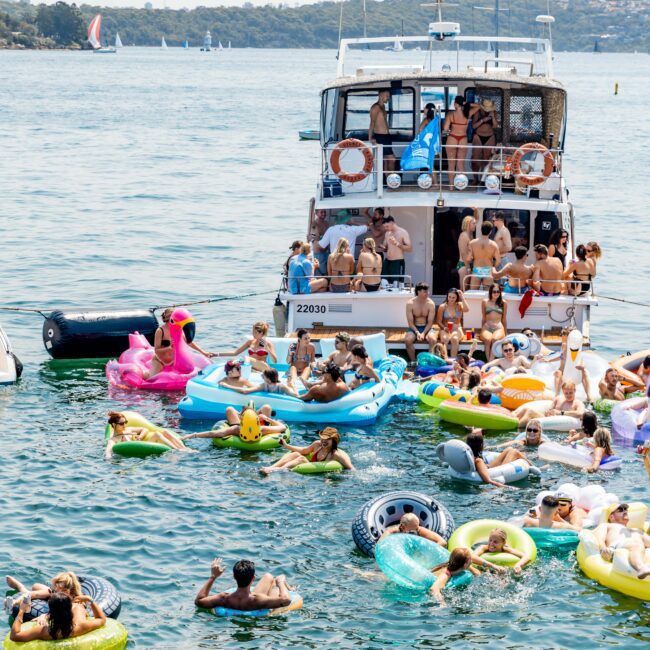 A lively boat party on a sunny day, with numerous people in swimwear floating on inflatable toys, including a pink flamingo and a rubber duck. The boat is anchored in the water with more people onboard, set against a backdrop of the coastline.