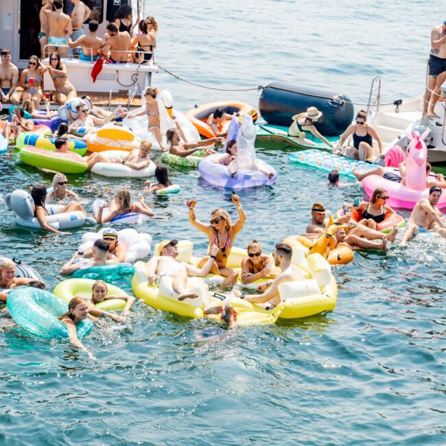 A group of people enjoying a sunny day on a lake, floating on colorful inflatable rafts and pool floats. There's a boat docked nearby with additional people on it. The scene is lively and festive, with clear blue water surrounding them.