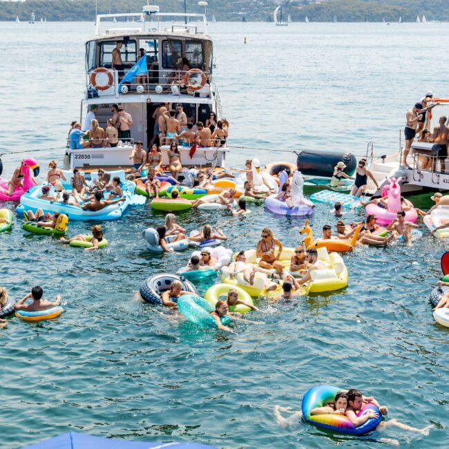 A lively scene of people enjoying a sunny day on a lake. Many are floating on colorful inflatable rafts shaped like animals. Boats are anchored nearby, and the lake is surrounded by forested hills in the background.