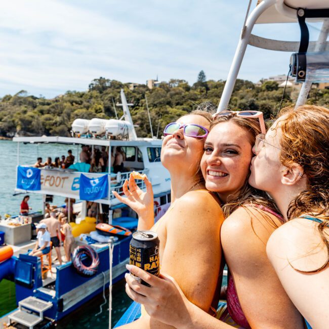 Three women pose happily for a photo on a boat, with one kissing another on the cheek. They are in swimsuits, and other boats and inflatable slides are visible in the background. It's a sunny day, and they seem to be enjoying a party or gathering.