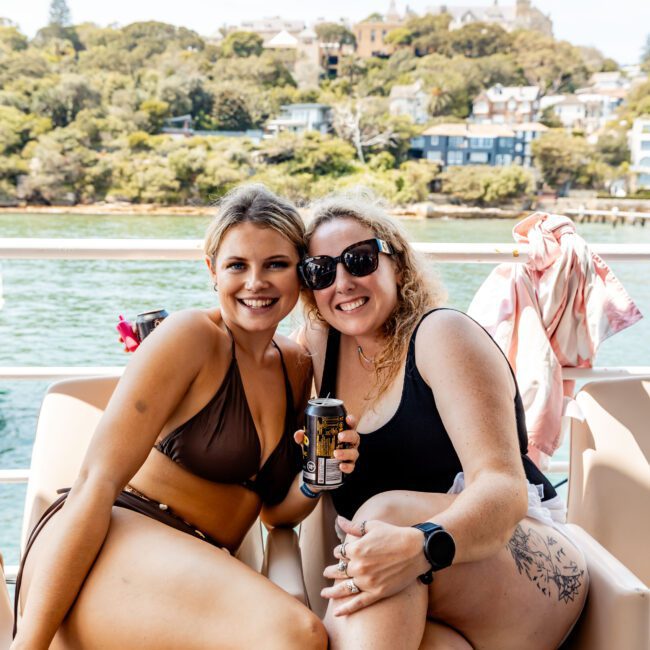 Two smiling women in swimsuits sit on a boat. One holds a drink can. There's a scenic view of water and lush greenery in the background, with houses visible on a hill.