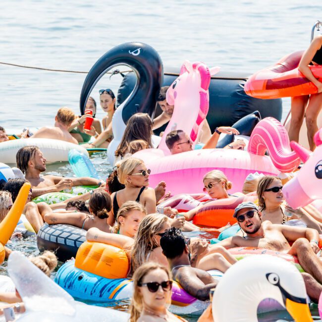 A large group of people enjoying a sunny day on a body of water, lounging on colorful inflatable floats shaped like animals. The scene is vibrant and lively, with a mix of swans, flamingos, and unicorns in various bright colors.