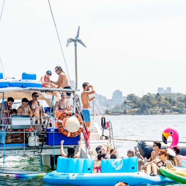 A lively scene with people enjoying a party on a boat and in the water. Several individuals are on inflatables, including a unicorn design. The city skyline is visible in the background under a clear sky.