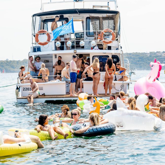 A group of people enjoying a sunny day on a yacht and in the water. Some are relaxing on inflatable pool floats shaped like a pink flamingo and a white swan. The atmosphere is lively and festive.