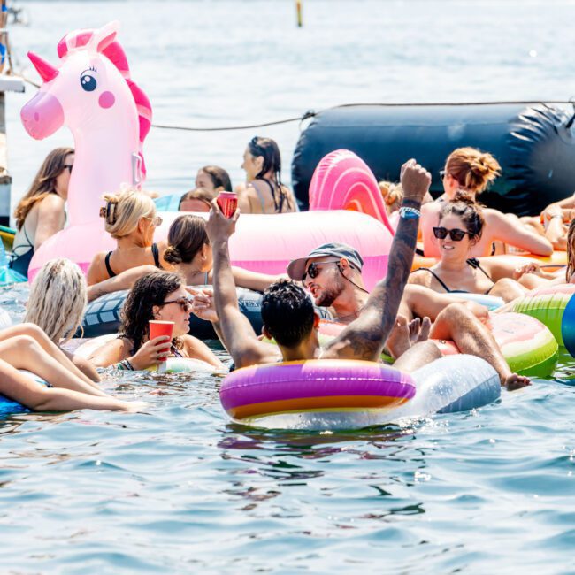 People enjoying a sunny day on a lake, floating on inflatables including a pink unicorn. Some hold drinks, while others relax in the water. There are hills in the background under a clear sky.