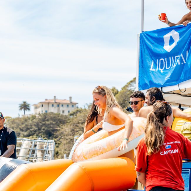 A woman wearing a bikini is about to go down an inflatable orange slide at a beach event. Several people are around her, including a person wearing a red shirt labeled "Captain." A building and trees are visible in the background.