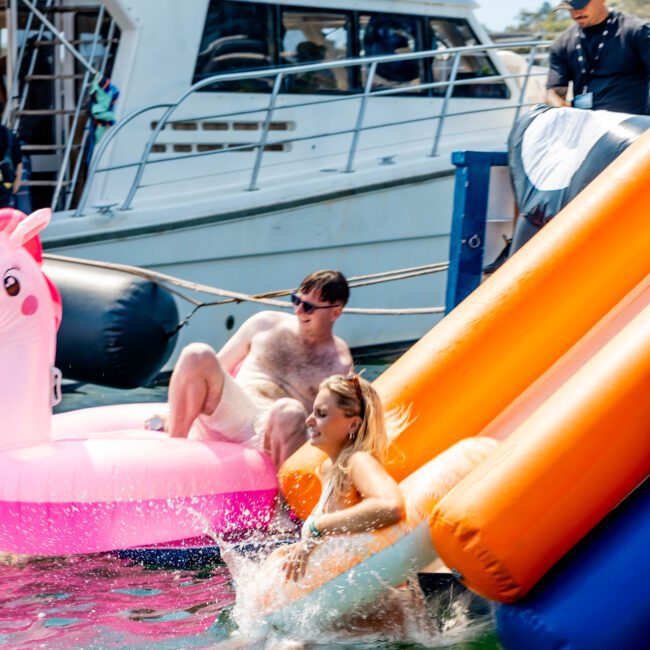 A woman splashes as she descends an inflatable slide into water. A man sits nearby on a pink float shaped like a unicorn. A boat is moored in the background with passengers on deck. It’s a sunny day at a social event on the water.