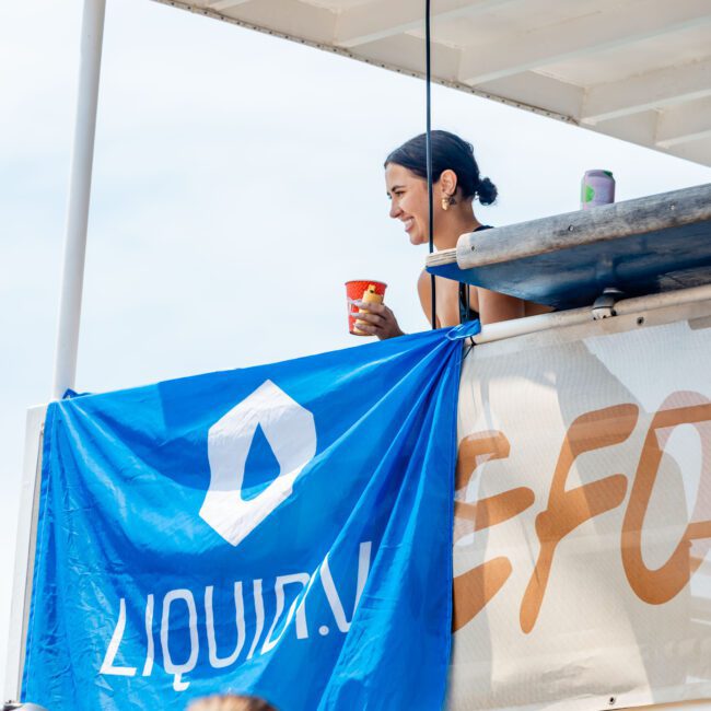 A woman stands on a boat's upper deck, smiling while holding a beverage. A blue "LIQUID IV" banner is draped over the railing. Other people are in the foreground, partially visible. The sky is clear and sunny.