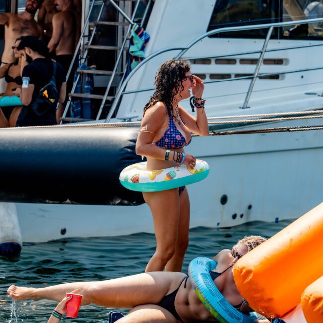 People are enjoying a sunny day on a floating platform by a boat. A woman stands with an inflatable ring, while others relax or swim. There are colorful inflatables and the water is calm.