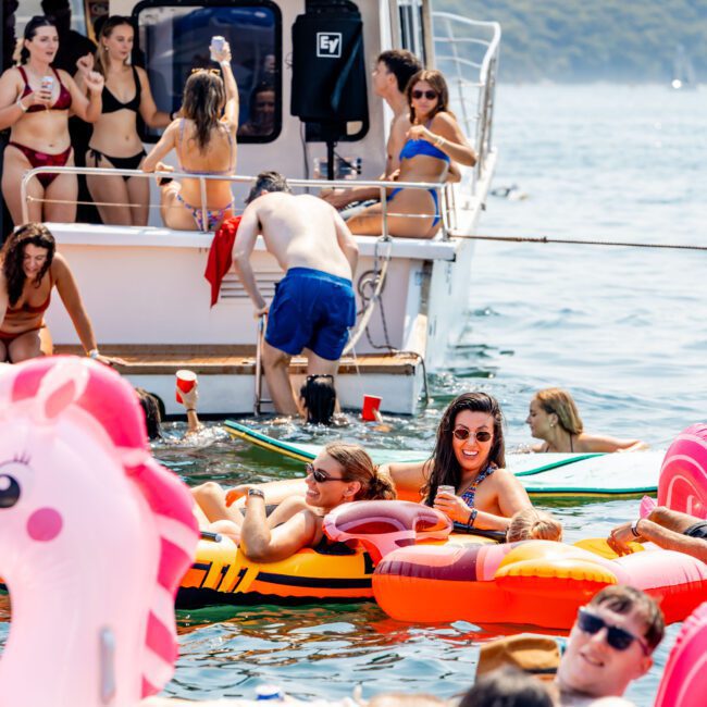 A group of people enjoy a sunny day on the water, floating on inflatable toys near a boat. Some relax on colorful inflatables like a pink unicorn and a doughnut, while others swim. The backdrop features lush green hills.