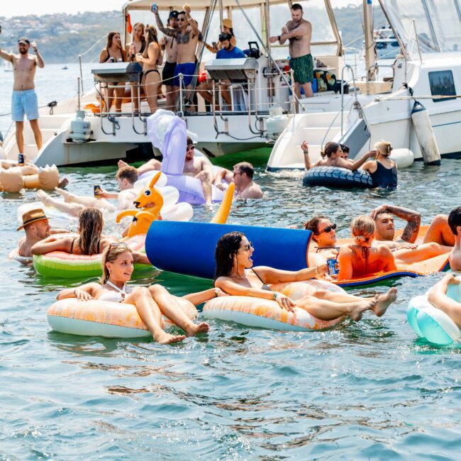 A group of people enjoying a sunny day on a lake, floating on colorful inflatables near anchored boats. Some are lounging on pool floats shaped like various animals while others stand or sit on the boats, taking in the lively atmosphere.