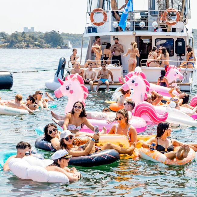 A sunny scene on a lake with a party happening on a boat. People are relaxing on colorful inflatable floaties, including unicorn and flamingo shapes, and socializing in the water. Music or conversation fills the lively atmosphere.