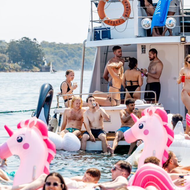 A group of people enjoying a sunny day on the water. They are gathered around a yacht, with some on the boat and others in the water on inflatable unicorns. The setting is lively and festive, with clear skies and trees in the background.