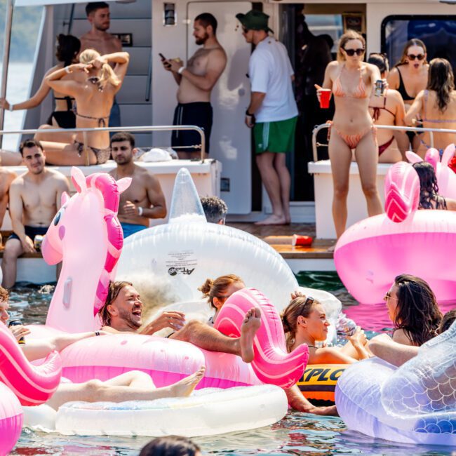 A lively boat party scene with people enjoying themselves on inflatable floats, including a unicorn and a shark. The boat's deck is crowded with partygoers, some holding drinks and chatting, while the water is filled with people relaxing and laughing.