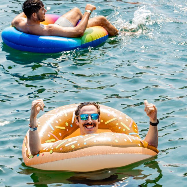 Two people float in a body of water on inflatable rings. One person is on a colorful raft, and another wears sunglasses on a donut-shaped float, smiling and giving thumbs up. Water ripples around them under a sunny sky.