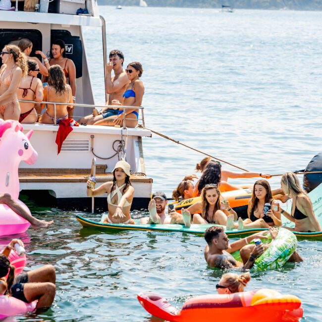 A group of people enjoy a sunny day on a boat and in the water. They are surrounded by colorful inflatable rafts and toys, such as a pink flamingo and an orange slice. The background features a calm lake and a forested shoreline.