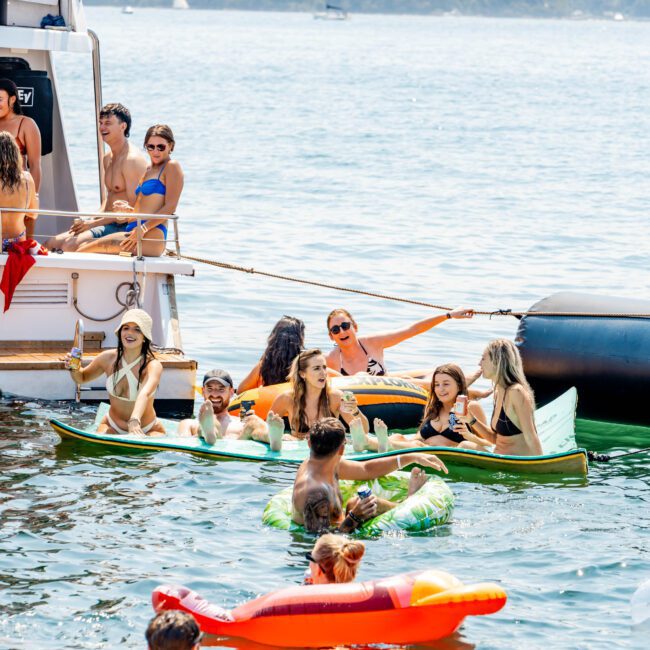 A group of people enjoying a sunny day on a lake, some sitting on a boat and others relaxing on inflatable floats shaped like an avocado and pizza. Trees and a shoreline are visible in the background.