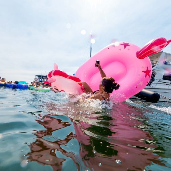 Person in a swimsuit sits on a large pink inflatable unicorn in the water, taking a selfie. Boats are in the background, and the scene is lively with people enjoying the water.
