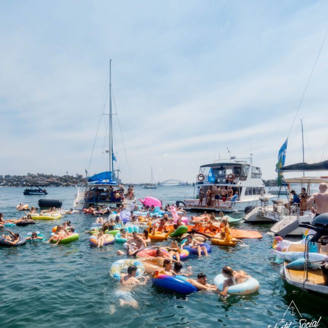 A group of people enjoying a sunny day on the water, floating on colorful inflatables around boats. The background shows a partially cloudy sky and a distant shoreline. The atmosphere is lively and festive.