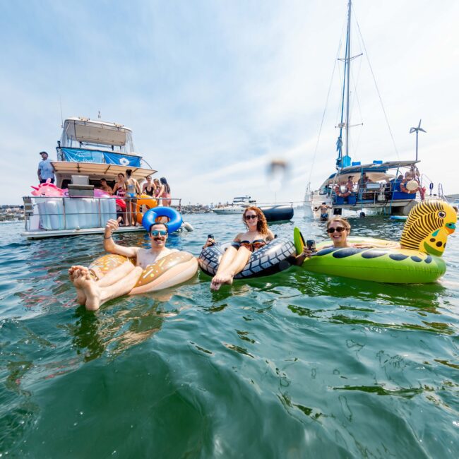 Four people are relaxing on inflatable floats in the water, with one in a pineapple-shaped float, another in a swan, a person on a dragon, and the fourth on a retro phone. A yacht and other boats are in the background on a sunny day.