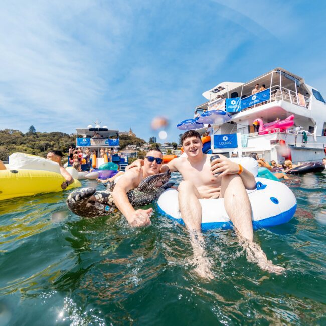 Two people in swimwear float on inflatable tubes in a sunny lake setting. They're smiling and posing for a selfie. A large boat and more inflatable rafts are in the background. Clear skies and a wooded shoreline frame the scene.