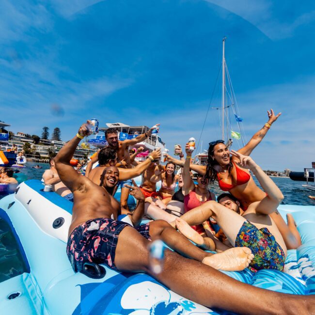 A group of people are enjoying a sunny day on an inflatable raft in the water. They are wearing swimsuits, laughing, and raising drinks. In the background, other people are on rafts and boats under a clear blue sky.