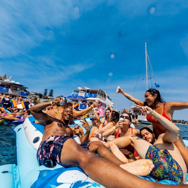 A group of people in swimwear are having fun on a large inflatable raft in the water, with boats visible in the background under a clear blue sky. They appear to be laughing and cheering.