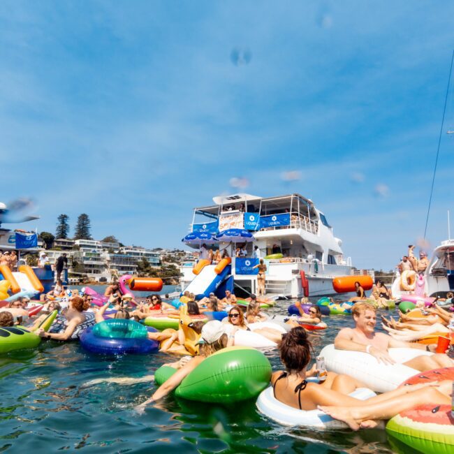 People are enjoying a sunny day on colorful inflatables in the water near a large yacht. The scene is lively and depicts a festive atmosphere with blue skies and coastal buildings in the background.
