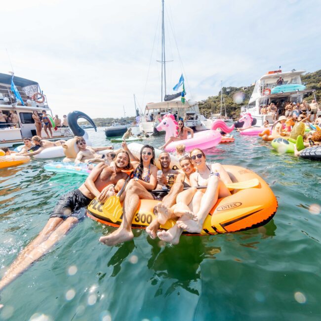 A group of people relax on inflatable rafts and floats in the water, surrounded by several boats. They're enjoying a sunny day, with some sitting on flamingo and pizza-shaped inflatables. Hills and trees are visible in the background.
