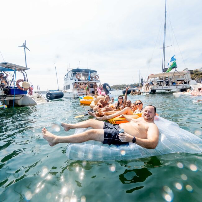 People are enjoying a sunny day on a lake, lounging on inflatable floats. Several boats are anchored nearby, and some people are swimming. The atmosphere is lively and festive.