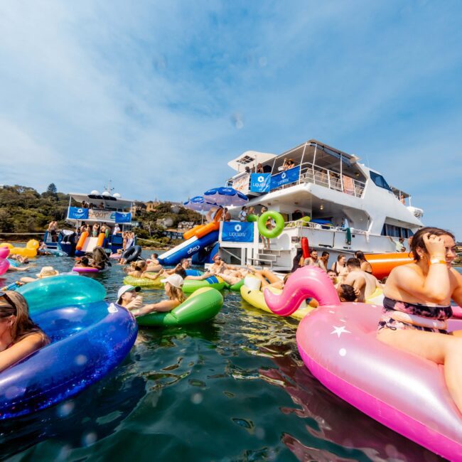 People in colorful inflatable rings float near a large white boat in sunny weather. The scene is lively, with various activities on board and in the water. Hills and trees are visible in the background under a clear blue sky.