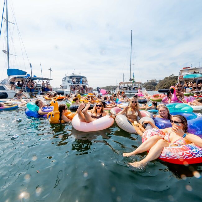 A group of people relax on colorful inflatable floats in the water near several yachts. The scene is festive and lively, with clear blue skies and a background of trees. The water is calm, and everyone appears to be enjoying the sunny day.