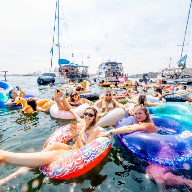 A group of people relaxing on colorful inflatable floats in the water, surrounded by yachts. The scene is festive, with individuals enjoying a sunny day, some wearing sunglasses. The background features several moored boats and a clear sky.