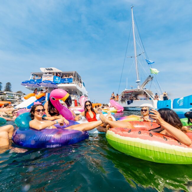 A group of people lounging on colorful inflatable floats in the water, near docked boats. The floats are shaped like a flamingo, watermelon, and other fun designs. The sky is clear and sunny, and partygoers appear relaxed and cheerful.