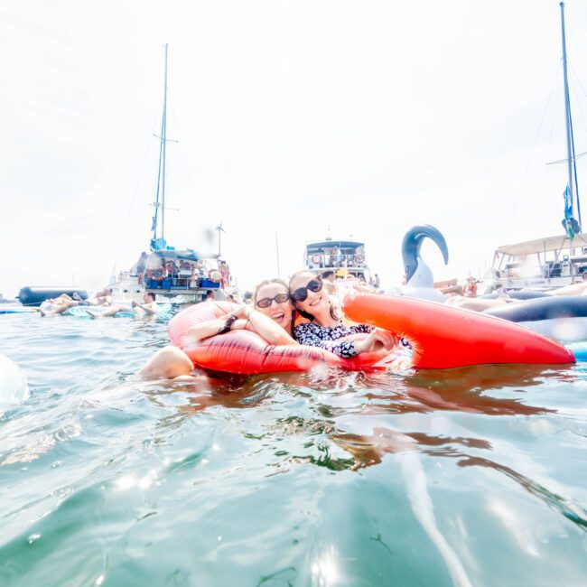 Two people are smiling and hugging while floating on a red inflatable ring in a body of water. Boats are visible in the background. The sky is clear, and the atmosphere is lively.