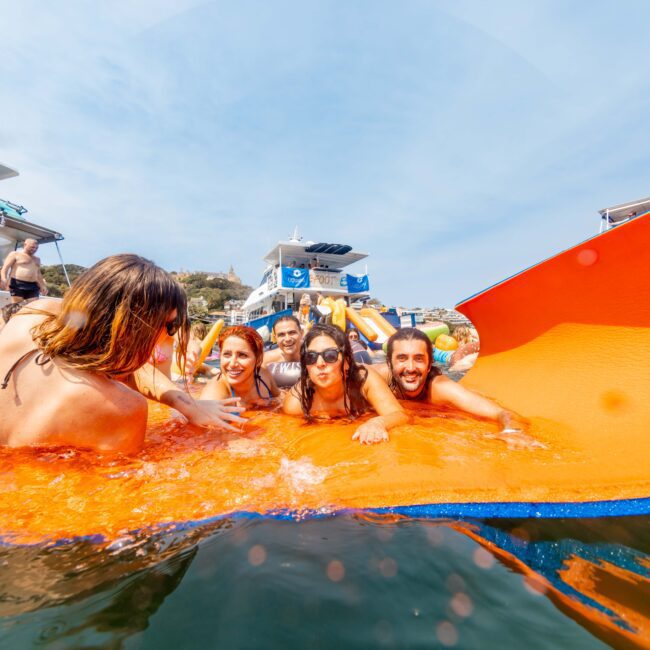 A group of people are joyfully lying on a large orange floating mat in the water near boats. They are smiling and enjoying a sunny day, with hills and blue sky in the background.