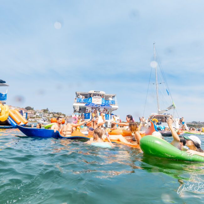 A group of people enjoying a sunny day on the water, using colorful inflatables, near a large boat. The sky is clear blue, and the scene looks lively and festive. Hills with buildings are visible in the background.