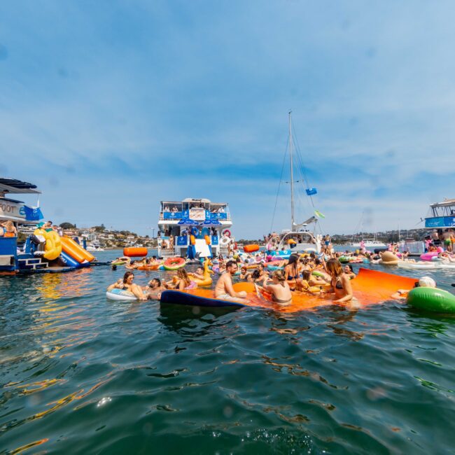 A vibrant gathering of people on the water, floating on colorful inflatables and enjoying the sunny day. Nearby, several boats are anchored, and a few flags are visible. The atmosphere is lively and festive, with clear blue skies above.