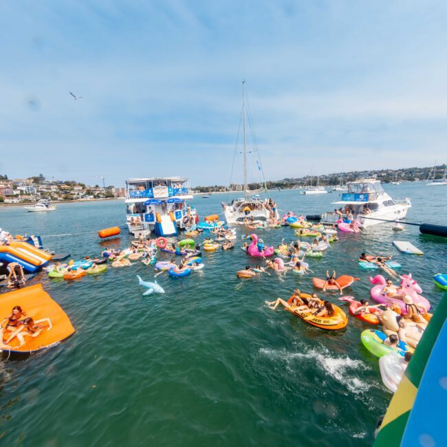 A lively scene on the water with people on colorful inflatable toys and boats. Several yachts and vessels are anchored nearby. The sky is clear, and there are buildings in the distant background. The image captures a festive, social atmosphere.