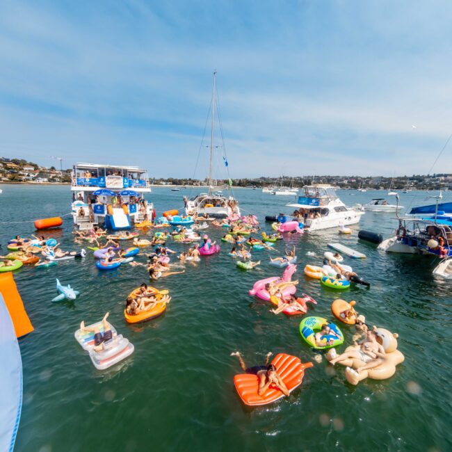A crowded marina scene with numerous people lounging on colorful inflatable floats in the water. Several boats and yachts are nearby, with a background of coastal houses and a clear blue sky.