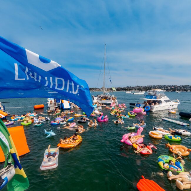 People enjoying a sunny day on a lake, floating on colorful inflatable rafts and toys. Boats are anchored nearby, and a large blue flag is waving prominently. The sky is clear, creating a festive and lively atmosphere.