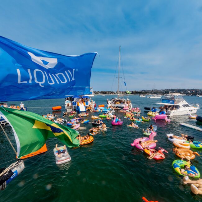 A large gathering of colorful inflatable rafts and floats are on a body of water, with boats anchored nearby. People are enjoying the sunny day. A prominent blue flag with "LIQUID" is in the foreground. The sky is clear and blue.
