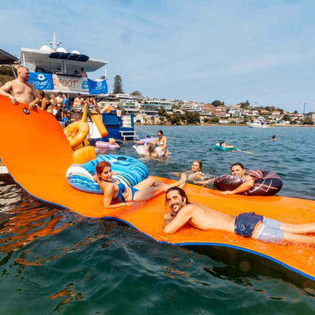 People are relaxing and having fun on an orange floating mat in the water. Some are on inflatable rings. A boat is anchored nearby, and there are buildings on the shore in the background. It's a sunny day.